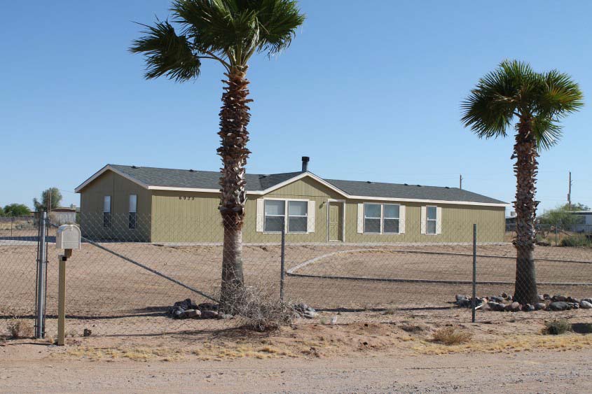 Green Mobile Home On A Dirt Lot With A Chain Link Fence Surrounding It.