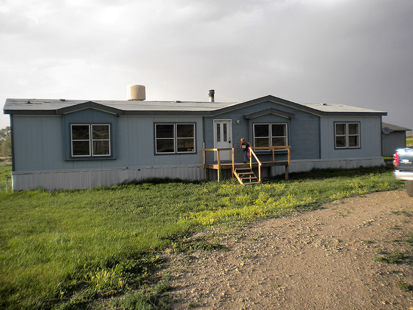 Slate Blue Mobile Home With A Dilapidated Porch And Broken Skirting.