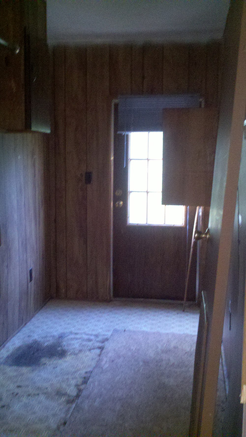 Small Room With Stained And Damaged Flooring, Wood Paneling On The Walls And A Door At The End.
