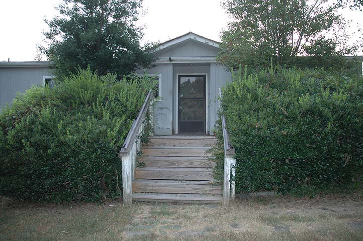 Grey Mobile Home With Overgrown Trees And Shrubs Growing Over The Edges Of The Damaged Porch.