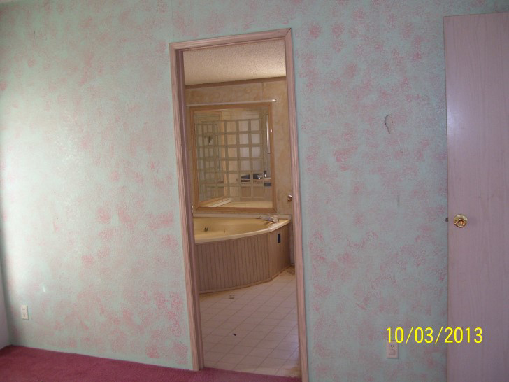 Bedroom With White And Pink Damaged Walls, With An Open Door Showing A Large Corner Tub.
