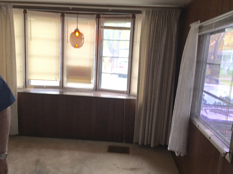 Room With Old White Carpeting And Aged Wood Paneling Below A Three Pane Window.