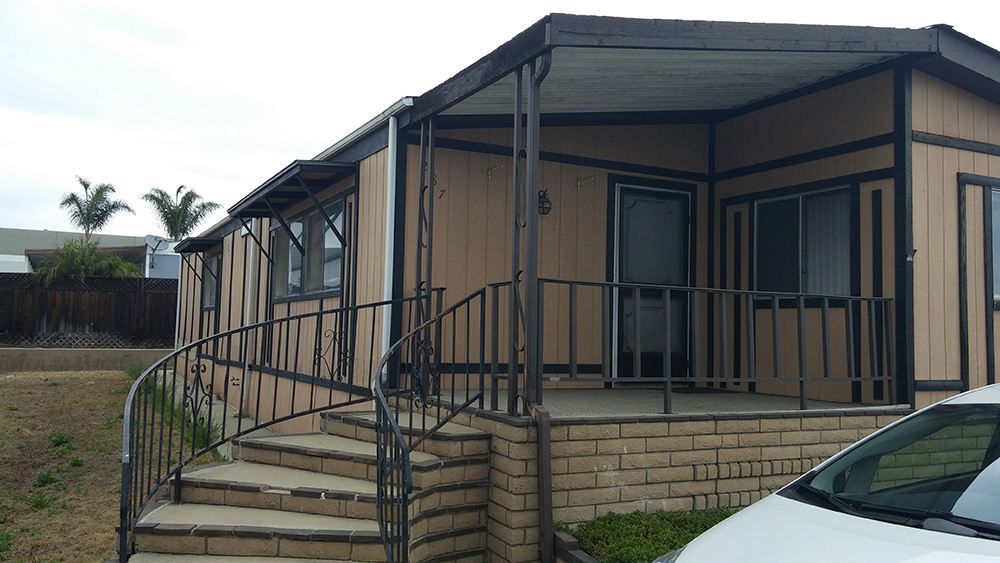 Outside Of A Brown With Black Accented Mobile Home With A Cement Front Porch And Curving Stairs.