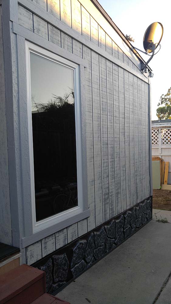 The Side Of A Mobile Home With Reil Rock Skirting and Distressed White And Grey Siding