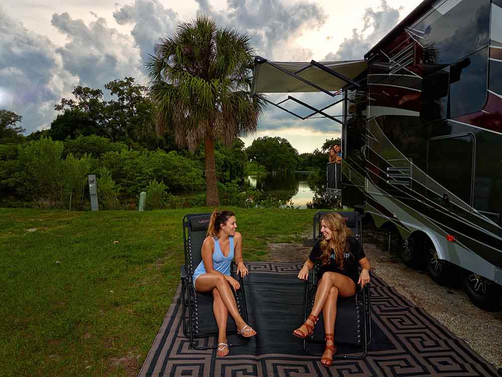 Two Women Sitting In Chairs Next TO An RV In Front Of A Lake.